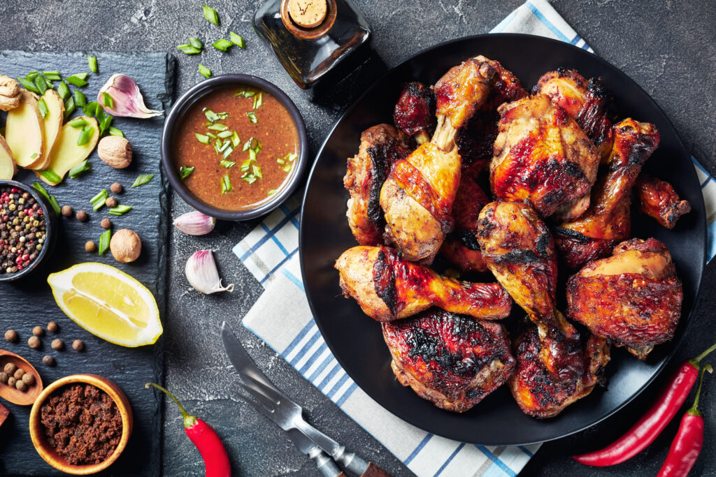 Spicy Grilled Caribbean Jerk Chicken drumsticks and thighs on a black platter on a wooden table with ingredients at the background, Anchorage Nutritionist, Jamaican recipe